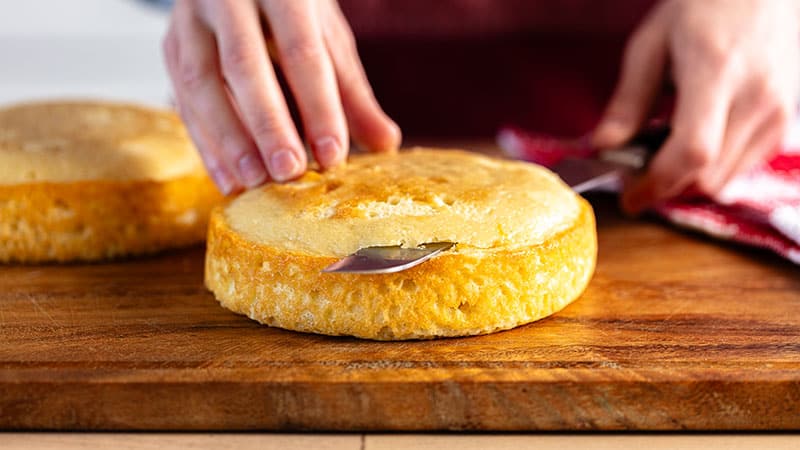 a person using a knife to trim yellow cake rounds on a wooden board&nbsp;