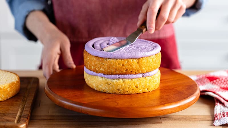 a person frosting a 2 layer cake on a wooden cake wheel&nbsp;