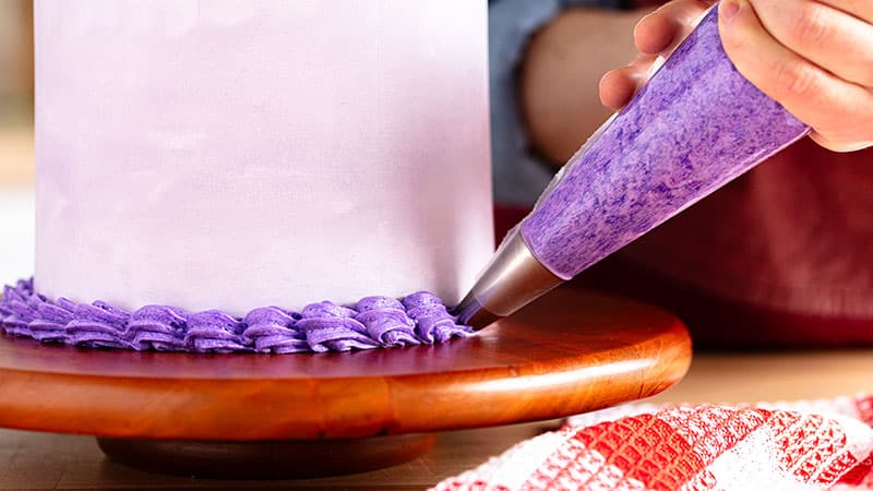a person pipes purple frosting shells around bottom of cake atop of wood cake wheel