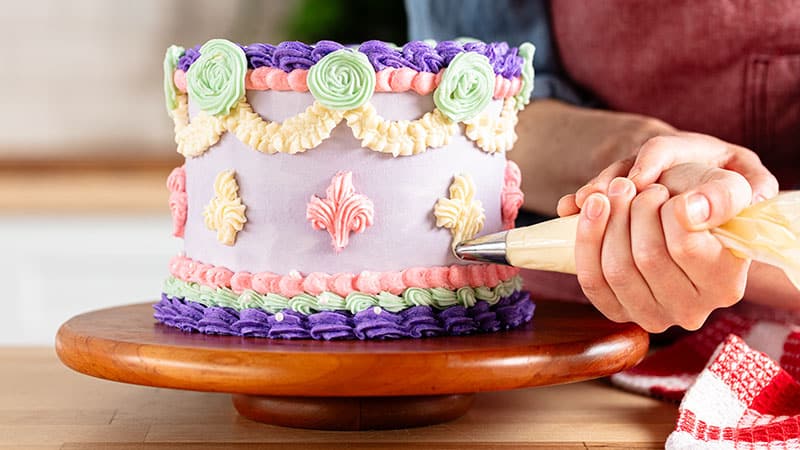 A person using a frosting bag to add fleu-de-lis designs to a cake