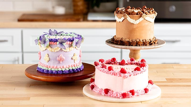 A wooden table featuring three cakes, each decorated with colorful frosting and intricate piping