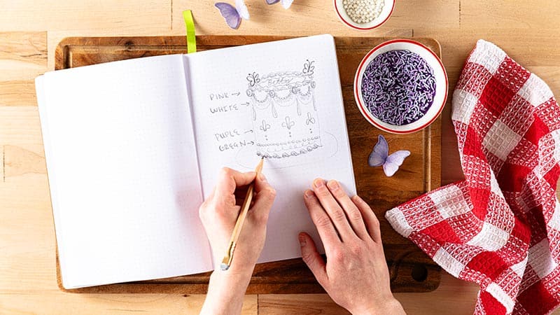 a person drawing a cake design onto paper, with bowls of decorations and a red and white napkin on the side