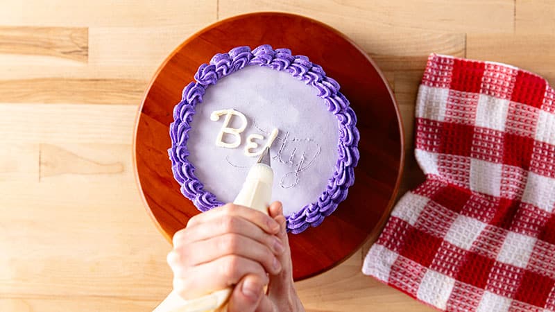  a person piping letters on top of a decorated cake&nbsp;