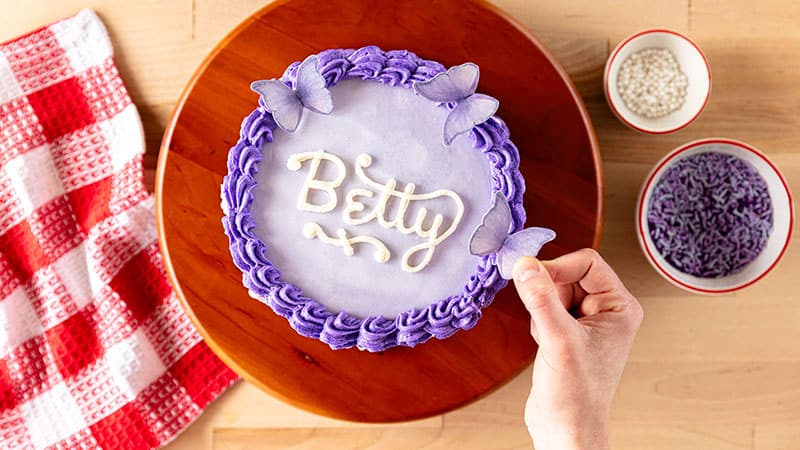 a person adding butterfly decorations to a frosted cake atop a cake wheel with bowls of sprinkles and red and white napkin on the side