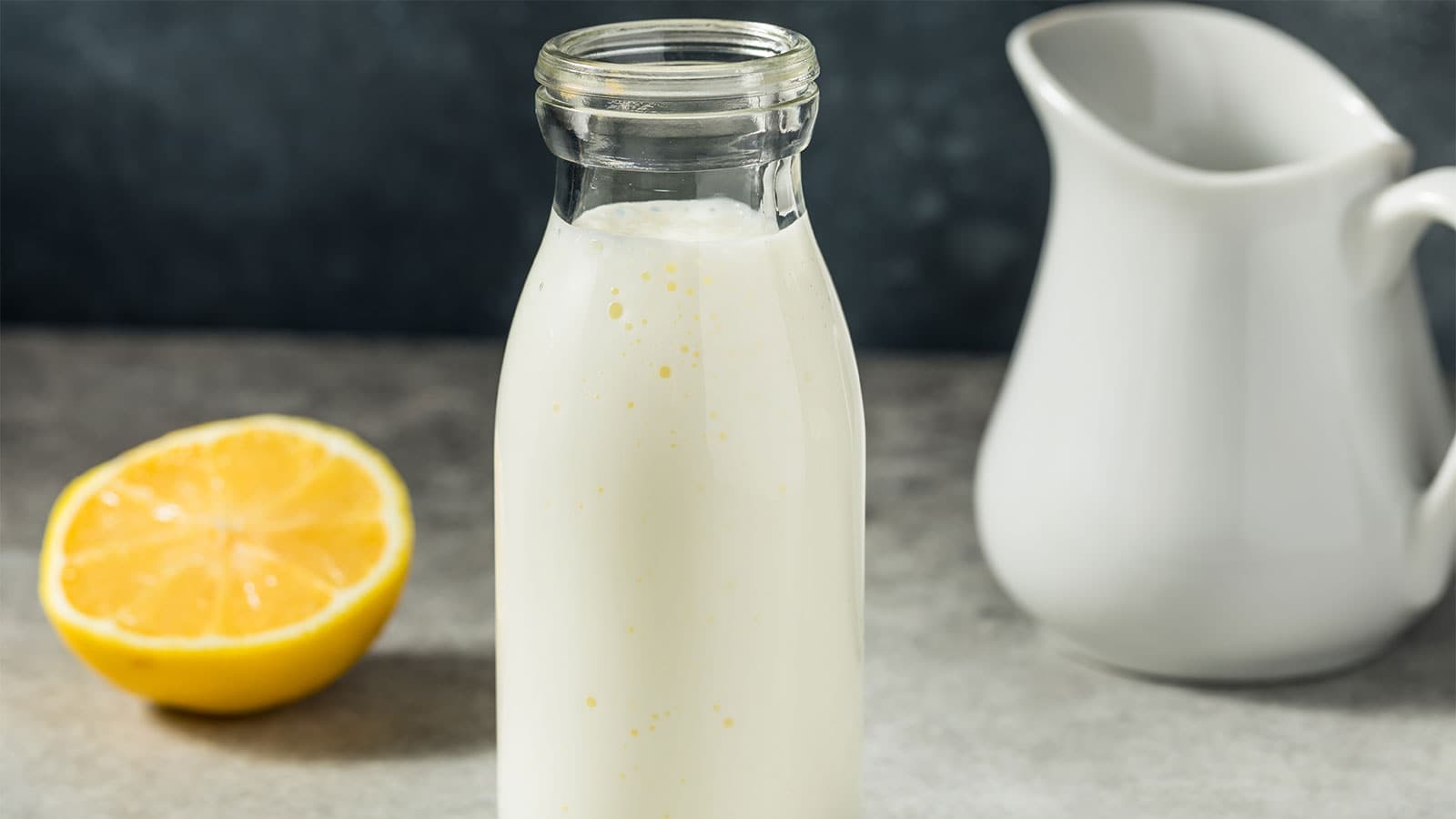 A glass bottle filled with buttermilk placed between a halved lemon and a white pitcher.