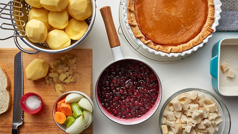 Peeled potatoes with a potato masher, a bowl of mirepoix vegetables, cranberry sauce in a pot, pumpkin pie, cubed bread in a bowl 